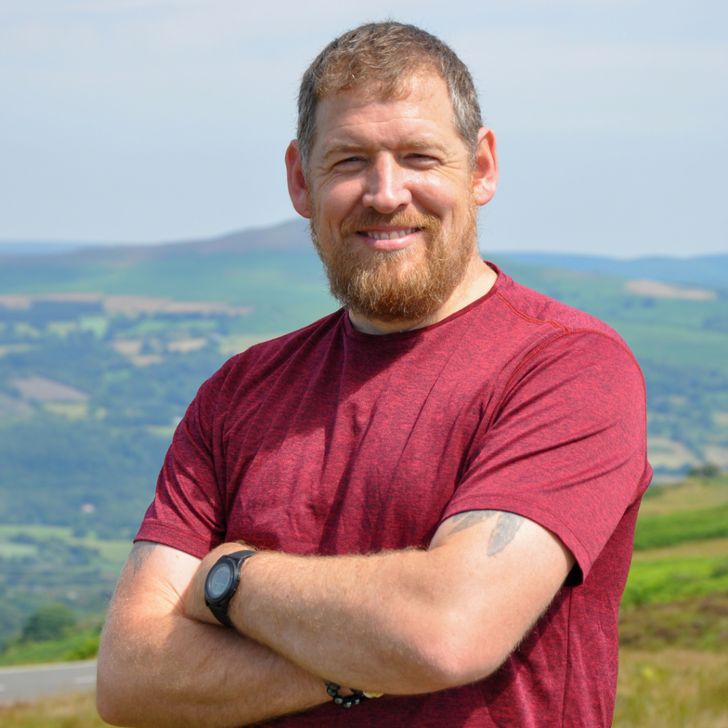 Jamie McAnsh smiling outdoors in a red shirt with arms crossed, standing against a scenic countryside backdrop.